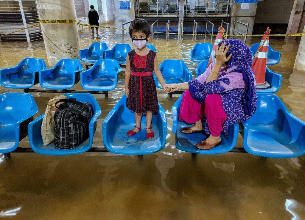 In the flooded lobby of Chittagong Maternal & Childcare Hospital in Bangladesh, a mother and her child, wait to receive medical treatment. They sit and stand on blue plastic chairs that appear to hover over the swirling, muddy water.