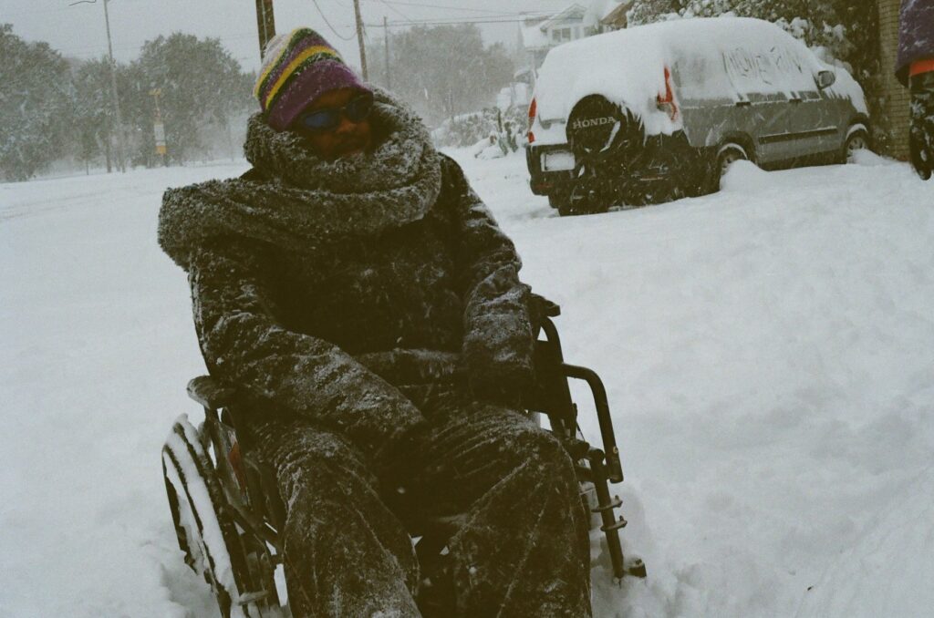 Captured on 35mm film during a historic Deep South snowstorm, this image shows Uhuru building a snow ma’am, reclaiming joy while reckoning with the inaccessibility of climate change. Snow packs into the wheels of his chair, a quiet reminder that extreme weather, made more frequent by climate crisis, further exposes the failures of ableist, racist, and transphobic public infrastructure. The grainy texture and subdued tones evoke a diaristic intimacy, grounding this moment of Black joy in a landscape shaped by both beauty and systemic neglect.