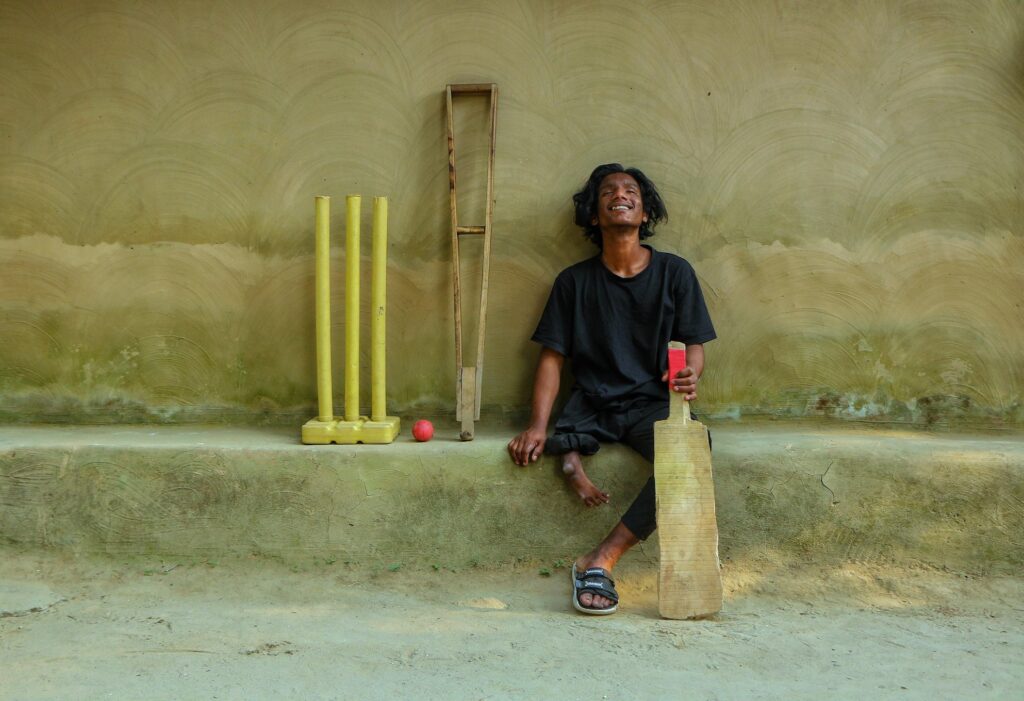 Jisad, a disabled cricketer born with a leg impairment, poses with his cricket bat, wicket, and crutch. His head tilted upward and a broad smile across his face, he radiates a spirit of determination and pride.Cricket has been Jisad’s passion since childhood—a love that brings him to life. Refusing to let his disability define him, he moves through the world with dignity and resilience. This portrait centres his joy, not his diagnosis, capturing an image of disabled life that is rarely foregrounded: one of passion, agency, and interiority.