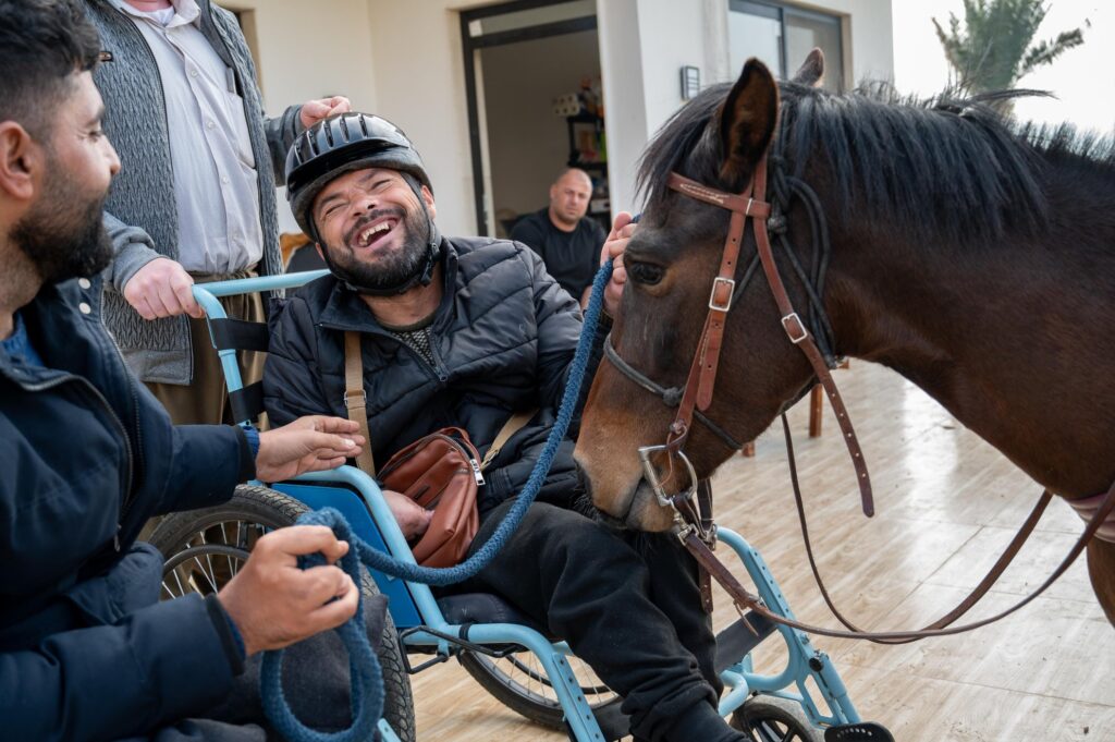 Salam Khalil, a Yazidi man, beams with joy as he interacts with a horse during a session at the Horses for Hope equine therapy project in the Kurdistan Region of Iraq. Though unable to walk, Salam’s radiant expression and open posture reveal a moment of unfiltered joy and connection—one that redefines what inclusive, therapeutic care can look like.This image underscores the power of intentionally designed spaces where disability is not a limitation but a starting point for reimagined forms of participation. Horses for Hope offers more than physical therapy; it cultivates dignity, belonging, and emotional restoration in a context where disability is often met with exclusion. Here, care is enacted not only by trained professionals but by peers, animals, and community members alike. The presence of men participating in this circle of care also gently challenges prevailing gender roles, expanding how we understand caregiving and emotional support. As a visual statement, the photograph insists on the centrality of both gender and disability in the creation of equitable and empowering environments.