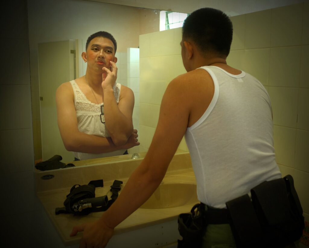 In a moment of quiet confrontation, a man gazes into the mirror- his stance grounded, his expression unreadable. On the sink lie his weapon and uniformed effects.