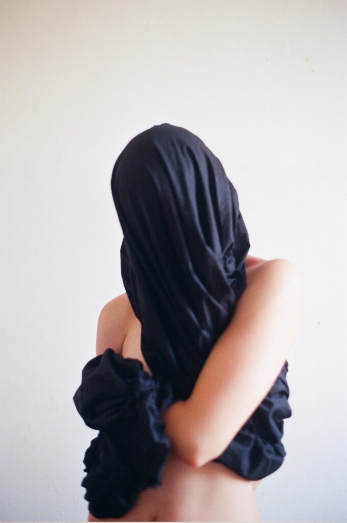 A figure stands facing the viewer, their head and chest wrapped tightly in black fabric, leaving only their belly, shoulders, and left upper arm exposed. The bright white background and stark lighting heighten the contrast between concealment and exposure, creating a powerful visual tension.Autism is an invisible yet deeply embodied disability that often goes unrecognised unless forcibly made legible to others. In this self-portrait, the artist refuses the demand for legibility. By concealing their face and upper body, they confront the viewer without capitulating to the expectation of visibility. This image documents a moment of sensory experimentation, where the body and the fabric become intertwined, existing beyond imposed meanings. The act of photographing becomes a form of empowerment, allowing the artist to assert their identity not as a fixed essence but as a dynamic, living being. Facing the camera is not necessary for recognition.As an autistic person, the artist often resists self-exposure, preferring a relationship with the camera and the world that privileges autonomy over forced disclosure. Through tactile and sensory engagement, they explore the intersections of gender, disability, and embodiment. This process, rather than the final image, is the heart of the work.