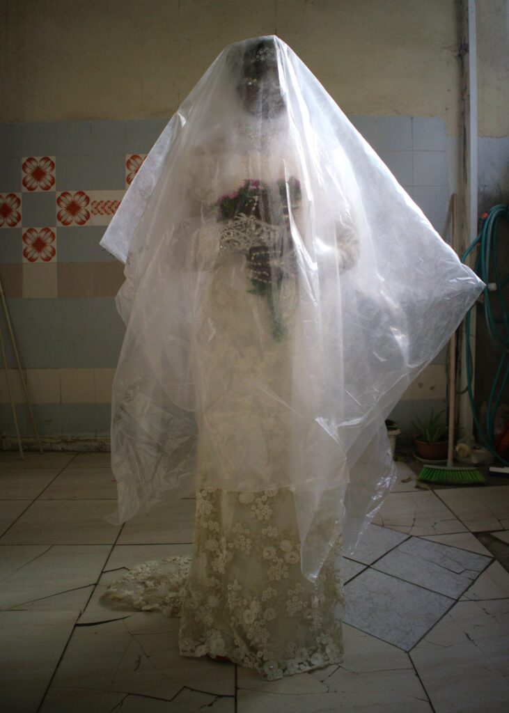 A Sudanese woman stands facing the camera, draped in a translucent plastic veil and holding a bouquet of white flowers. Her stance and attire evoke a bridal aesthetic—but the materials and context disrupt any sense of celebration. The veil suggests not just ceremony, but also containment, displacement, or medical control.This image is part of The House is Black, a performative photography project exploring the intersection of gender, disability, and migration through the lived experience of Hawa, a Sudanese asylum seeker and single mother in Tel Aviv. Inspired by Forough Farrokhzad’s 1963 film of the same name, which documented life in a leper colony, the series uses poetic and symbolic language to foreground often-unseen realities. Working collaboratively with the artist, Hawa becomes both subject and storyteller. The project challenges dominant portrayals of refugee women, shifting the focus from passive victimhood to the complex labour of caregiving, survival, and resistance. It draws attention to invisible disabilities—both the physical toll of care work and the emotional burden of displacement—and demands a deeper reckoning with how gender, health, and migration intersect.