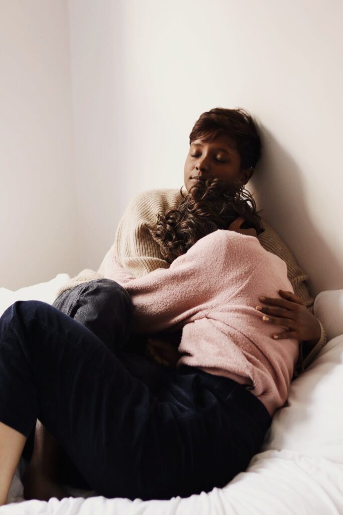 Kai and Irene sit closely together, enveloped in a deep embrace. Irene presses their face into Kai’s chest, who cradles them with steady, protective care. Their bodies fold into each other, creating a powerful sense of intimacy, trust, and grounding.