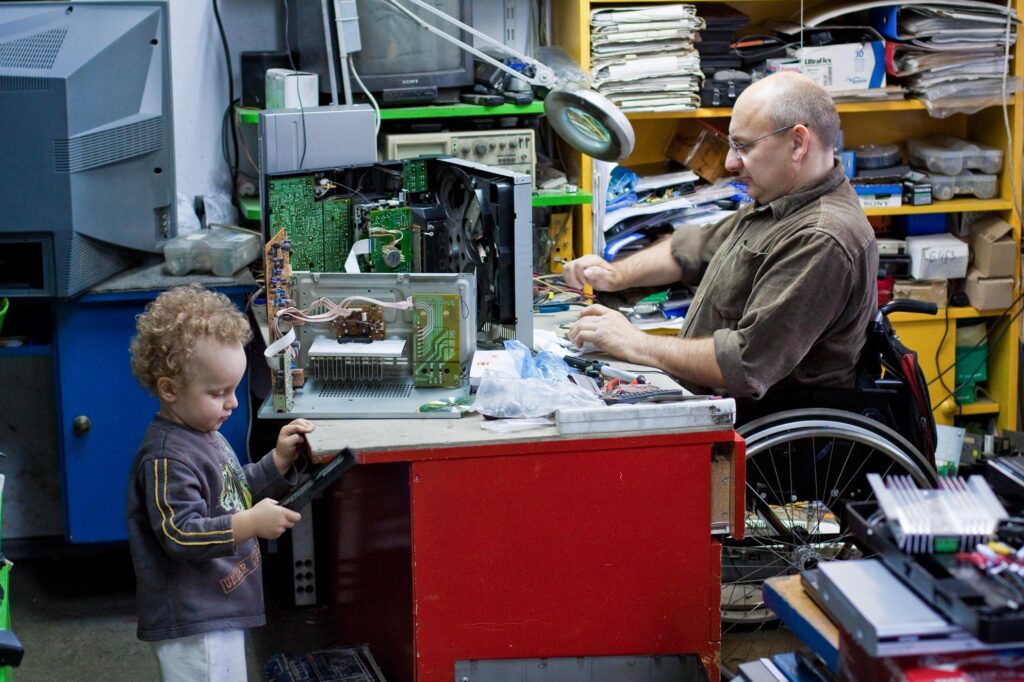 A father works at his cluttered desk from his wheelchair, focused and composed, while his young son plays nearby. The scene is filled with quiet, everyday warmth, a depiction of work, parenthood, and care unfolding side by side.