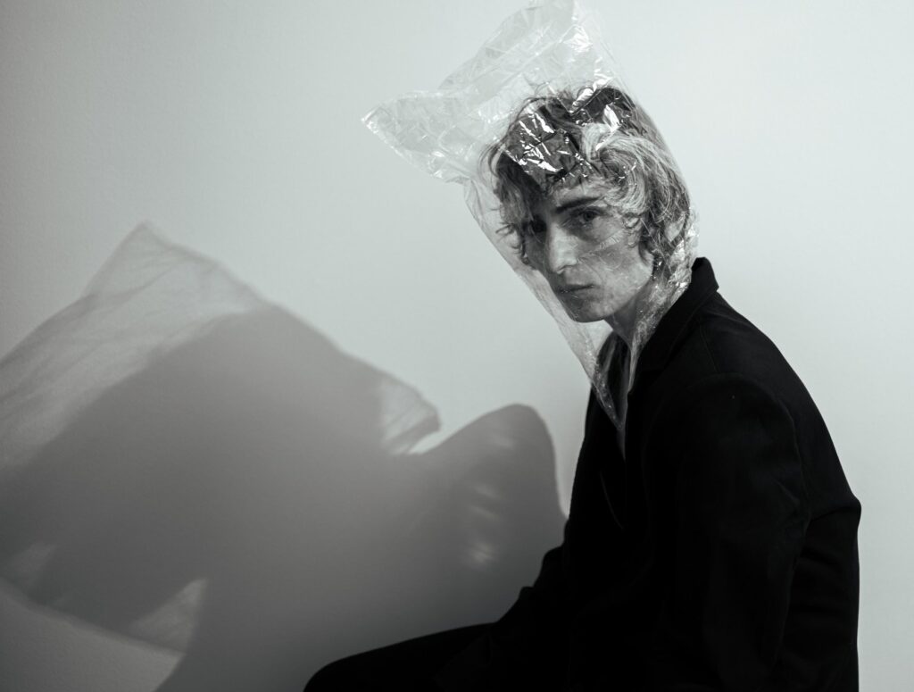 A figure confronts the camera head-on, their gaze unwavering. Over their head, a clear plastic bag, at once suffocating and delicate, like a veil fashioned from constraint. The play of shadow distorts the body behind them, casting an echo of their form that is both haunting and poetic.