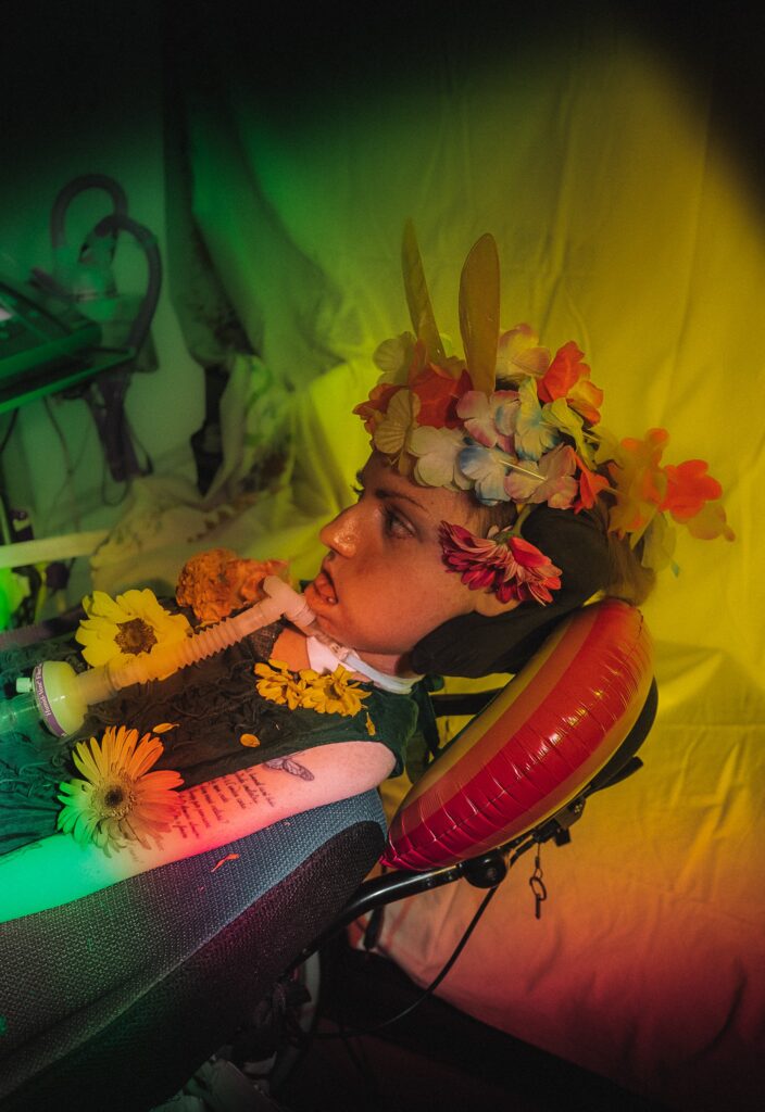 A young girl lies in bed, her breathing apparatus and elaborate floral headdress entwined in a dreamlike display. Bathed in multicoloured light, she gazes away from the camera, her expression distant and absorbed, as if looking beyond our reach.
