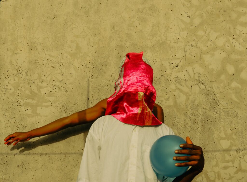 A male subject poses against the backdrop of a textured wall, his face obscured by a vibrant red fabric. He cradles a fragile blue balloon in one hand, while an arm, delicate and feminine, extends behind him, reaching outward.