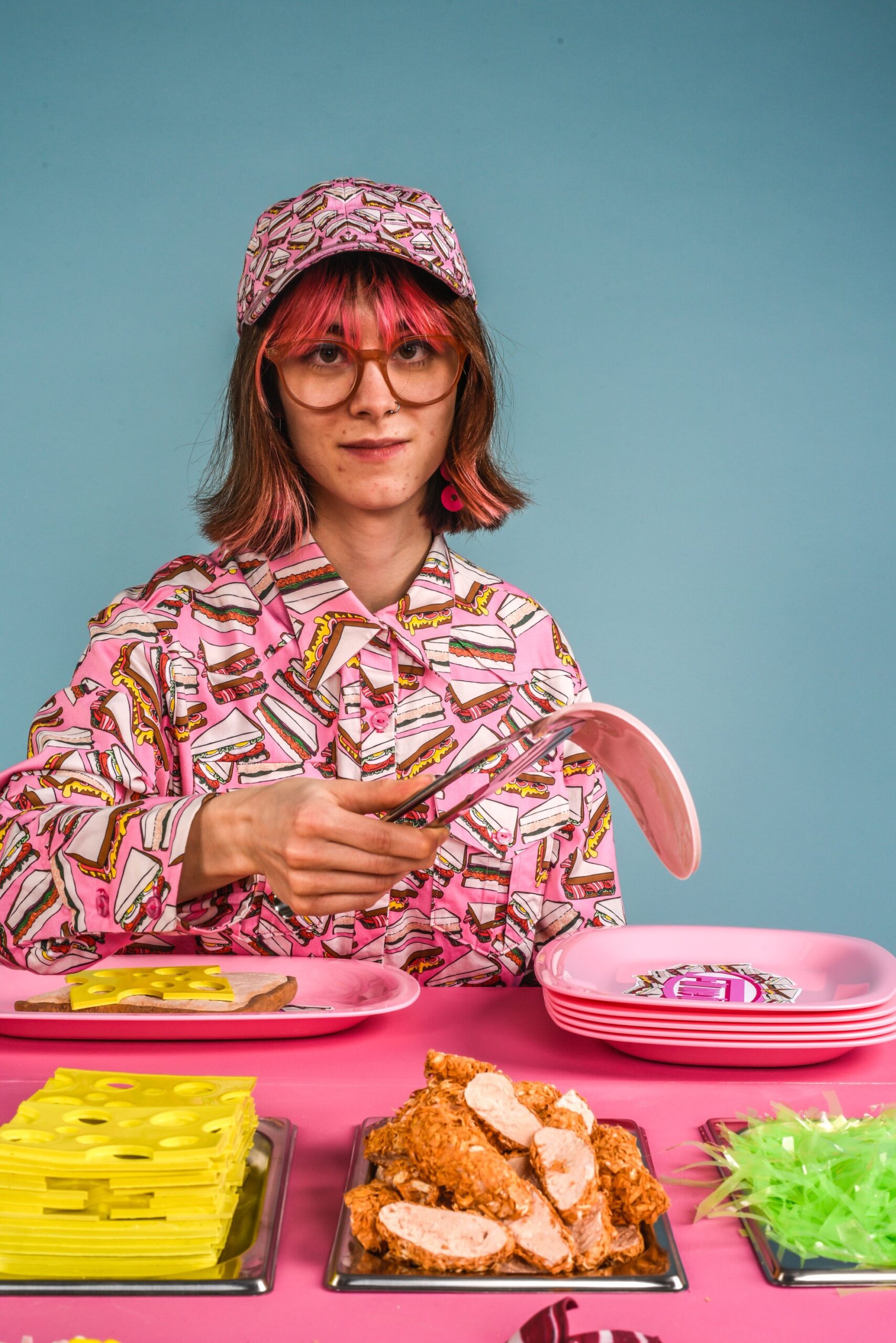 Dressed in a bright pink sandwich shirt and hat, Milly prepares a sandwich in this surreal, playful installation. Plastic cheese, bread, salad, and meat evoke both childhood imagination and a deeper commentary on exclusion.