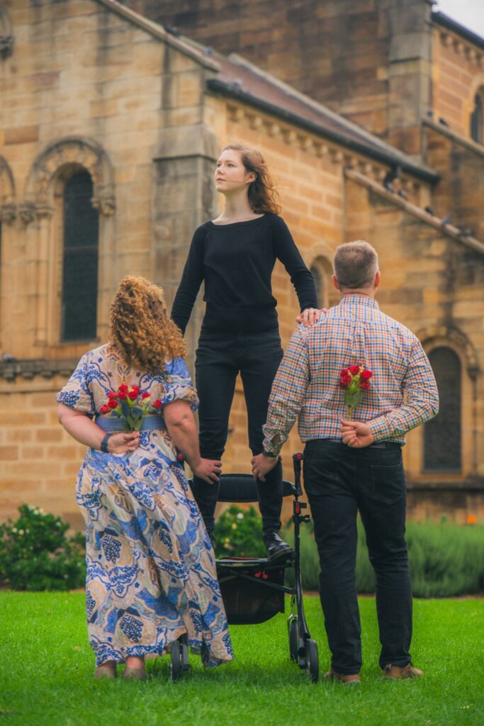 Macy is lifted high by two loved ones, their body framed against open sky. The gesture is one of intimacy and strength—a moment of vulnerability met with unwavering support.