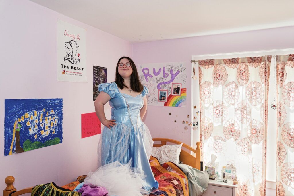 Ruby Kaftan, 20, poses confidently in her bedroom wearing her favourite blue princess dress. Her hands on her hip, she radiates self-assurance and agency. A writer, illustrator, and fashion designer, Ruby uses creativity to shape a world that reflects her identity on her own terms.
