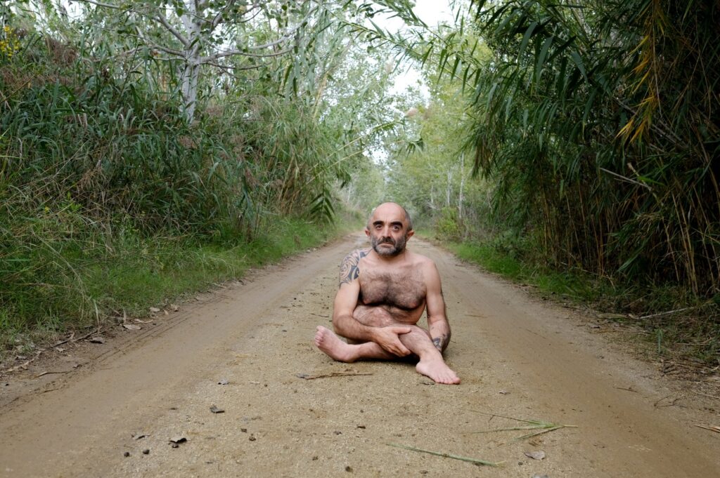Seated at the centre of a long, straight road, Íñigo meets the camera’s gaze head-on. Flanked by dense greenery, his presence is grounded, quiet, and resolute.