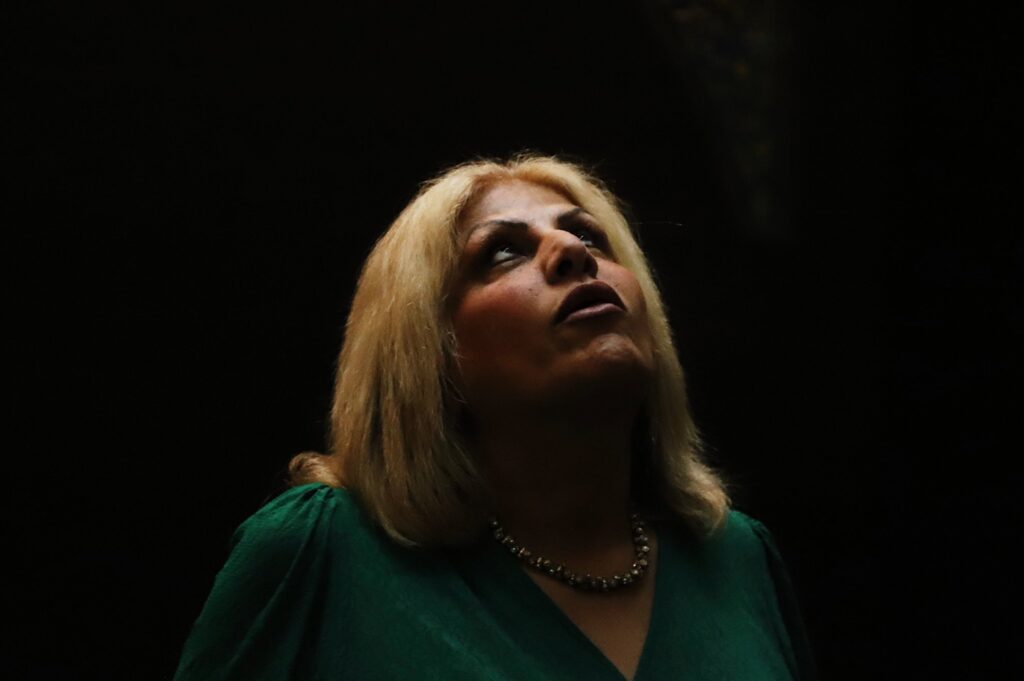 Bathed in a solitary spotlight against a sea of darkness, Fereshteh gazes upward, her mouth gently pursed as if in mid-song or suspended breath. Her expression holds a poignant tension, between longing and proclamation, between silence and sound.