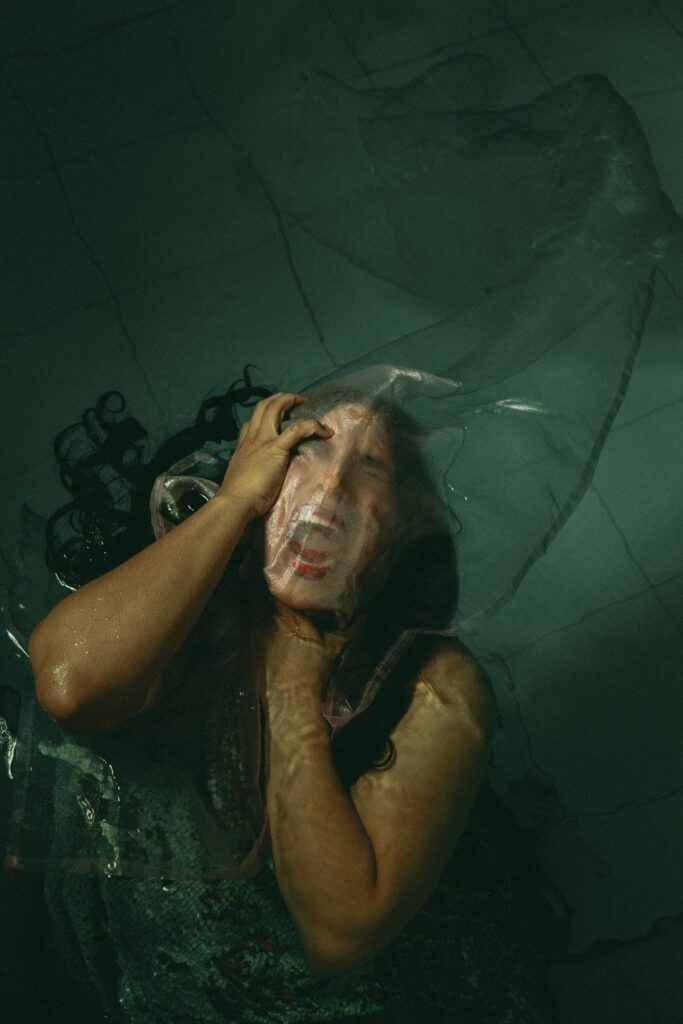 A person screams, her hands clutching her neck, her face encased in a fabric bag. Here, the murky, dark hue of the background and contorted gestures evoke a visceral sense of pain, trauma, and suffocation.