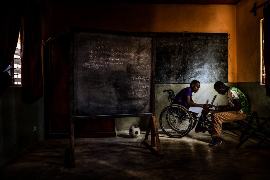 Kodjo, a 14-year-old boy in a wheelchair, sits with quiet purpose in a makeshift classroom at the Orione Center for children with disabilities. Caught mid-action as he assists a peer with homework, his presence radiates a calm, pedagogical authority. The composition, intimate and gently distanced, offers the viewer an unstaged glimpse into a moment of mutual care and solidarity.