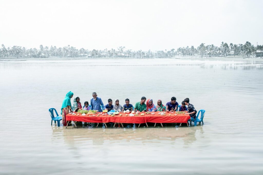 On an island bordering the Sundarbans Forest in Bangladesh, four fishermen’s families gather around an orange table, their remaining food supplies set before them. Blue plastic chairs sink into ankle-deep water. While they appear vibrant in the foreground, the surrounding landscape fades into a grey haze of distant trees and land.