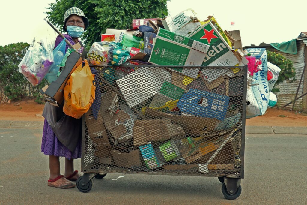 Gogo Mary Mauze poses with her recycling cart. Her gaze meets ours, face partly obscured by a blue medical mask. Ankles swollen in worn slippers, she appears wearied yet unyielding.