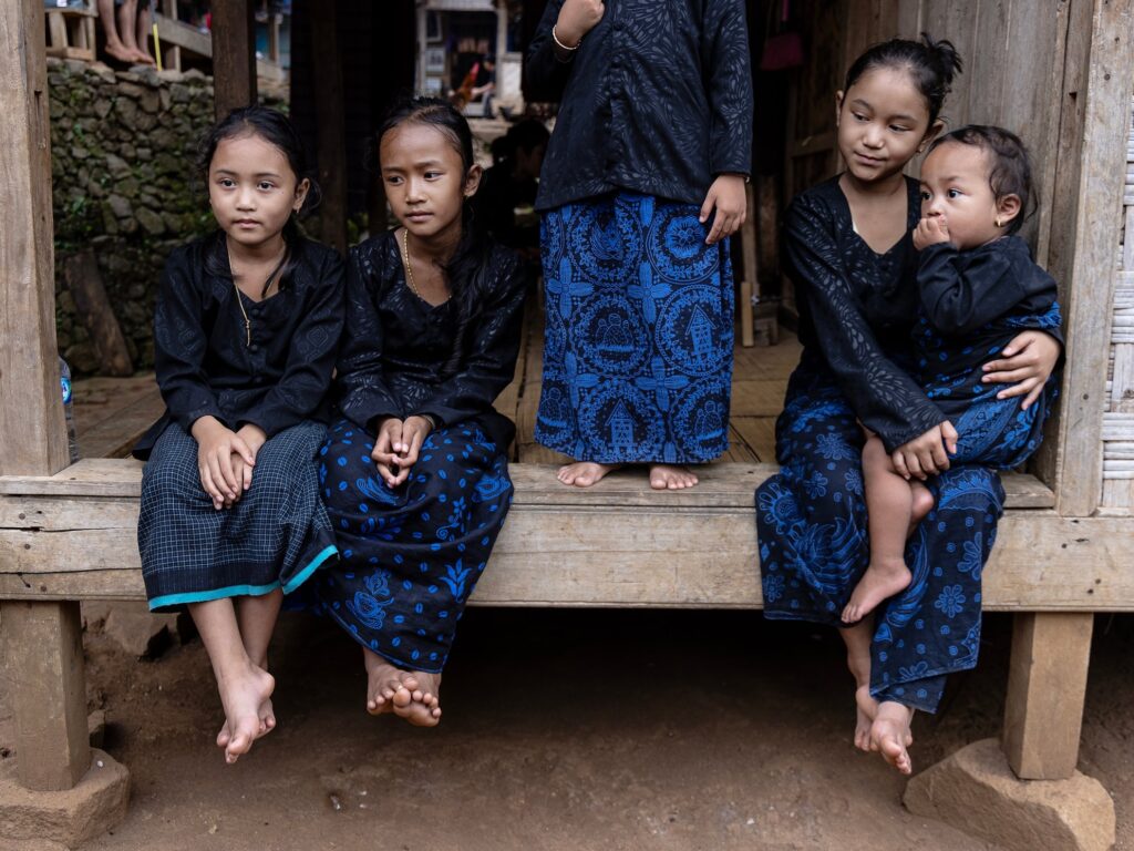 Dressed in black and blue kebaya, four young girls and a baby sit on the steps of their home in a Baduy village. Legs swinging, hands folded in their laps, one girl cradles the infant while another stands watch.