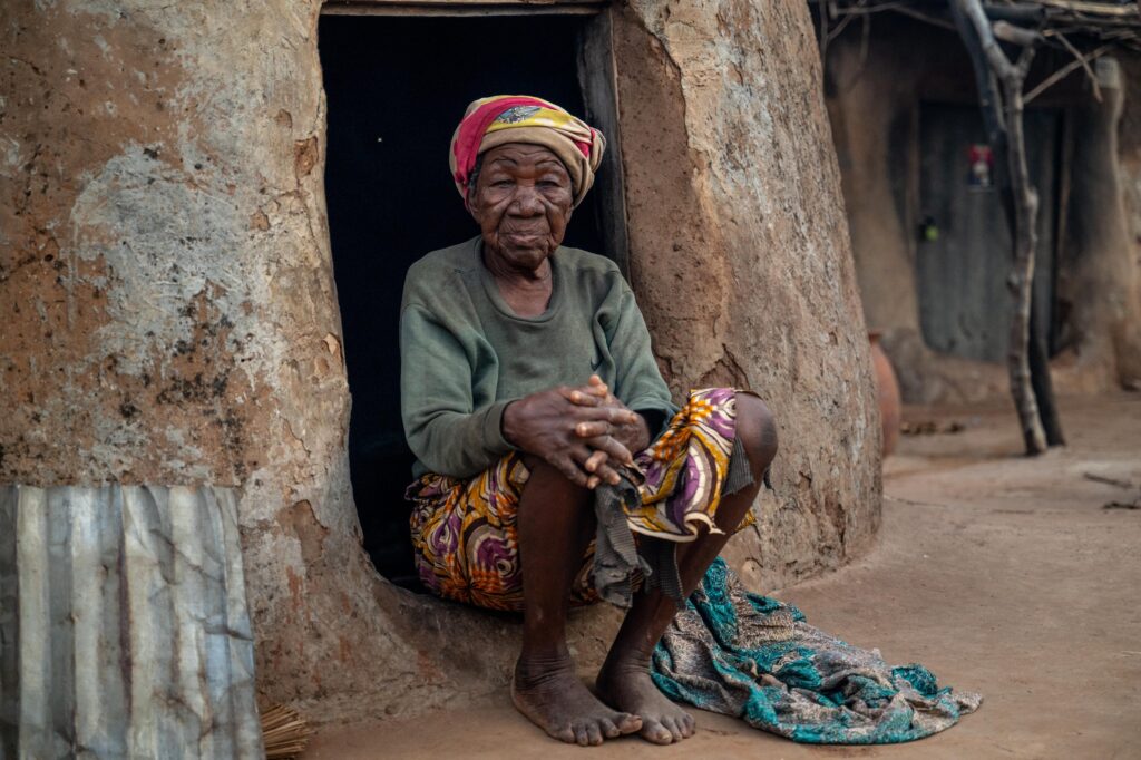 Bachalbanueya sits at the entrance of her home in the Gambaga camp, northern Ghana, where she has lived in exile for over forty years. Hands resting on her knees, she meets our gaze directly.