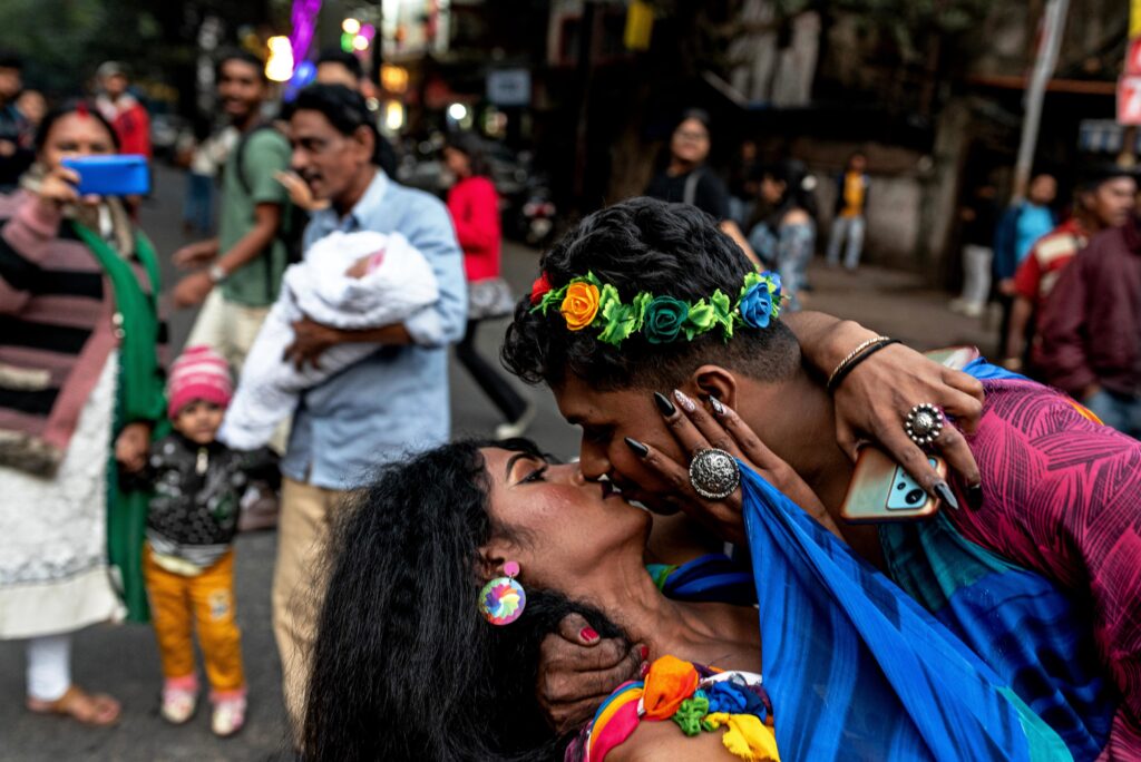 A transgender couple kisses during a Pride Parade in Kolkata, their embrace a public claim to love and dignity. Behind them, a young family smiles and photographs the moment.
