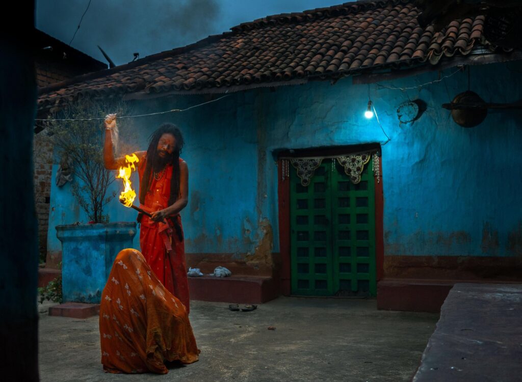 Padma Shri Chutni Mahato sits with her knees drawn to her chest as a tantrik performs a ritual before her, a torch of fire raised high. Behind them, the faded blue of a village building sets the scene — ordinary yet charged. The confrontation feels both intimate and symbolic: two belief systems facing one another, one wielding fear, the other quiet defiance.