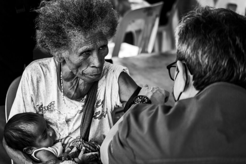 An elderly woman cradles a sleeping baby as a healthcare worker takes her blood pressure. Rendered in black and white, the scene balances intimacy with confrontation.
