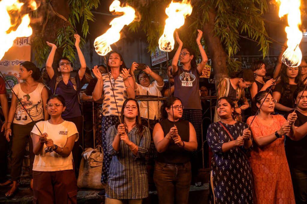 A group of women chant and clap, lit by the glow of burning torches. Arms raised mid-air, gathered close together, they appear resolute as the flames flicker high against the night.