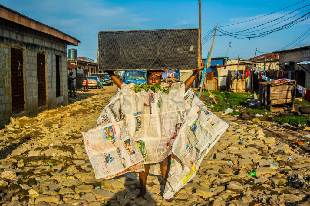 A woman stands defiant amid the detritus of Lagos, dressed in a gown made of newspapers and holding a large speaker above her head. Leaves fill her mouth, as though speech itself has become elemental. Staged in the Iwaya community, the image transforms protest into performance — media, nature, and sound converging in an act of resistance.