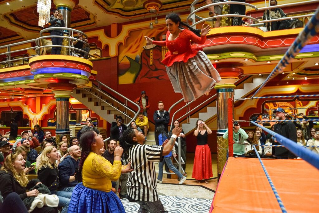 Benita launches from one of the ring’s poles, mid-air above her opponent and the referee, as the crowd erupts in awe. Suspended mid-leap, the scene is charged with colour and momentum, embodying a radical reclaiming of justice, performed, celebrated, and shared with an audience.