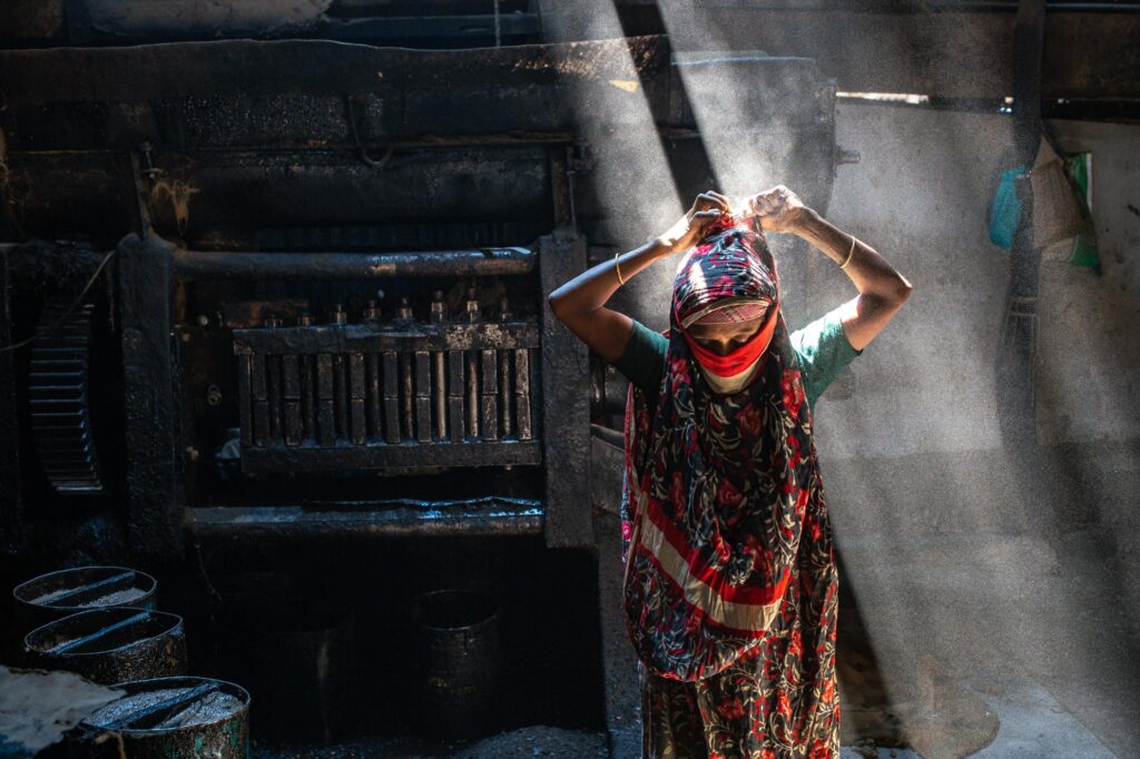 Rahima Khatun stands in the half-light of a smoke-filled room, her hands raised above her head, bracelets glinting as if caught between labour and restraint. The shaft of light that falls across her body cuts through the dimness like bars — a carceral echo rendered in shadow and air.