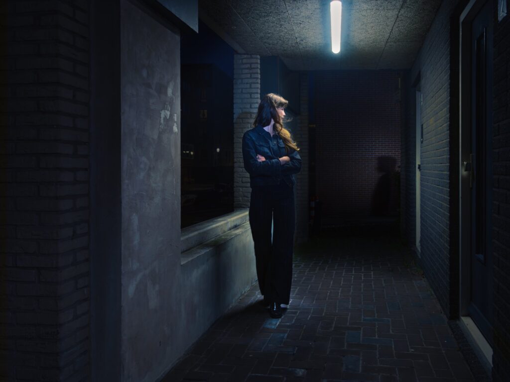 A woman stands in a dim, narrow corridor at night. Her posture is taut as she turns back to the encroaching shadows. A single overhead light casts sharp contrasts; in the corner, another figure emerges, indistinct yet present. The corridor closes in, suffused with claustrophobic unease.