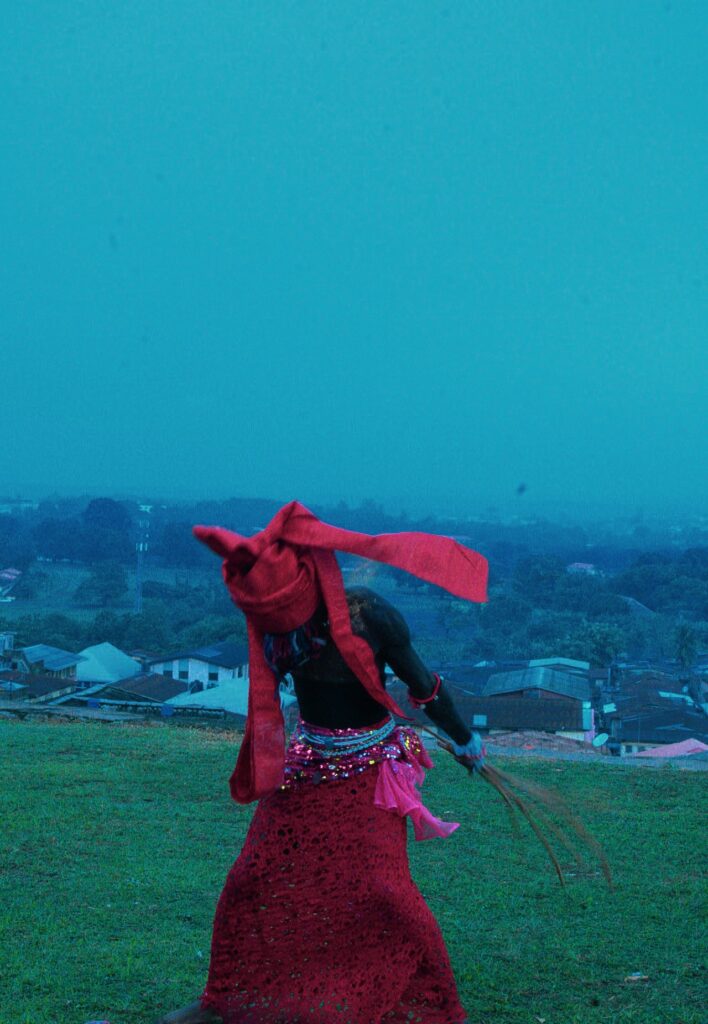 Egbiameje Omole performs Pankere in Glitter, a work confronting the traumatic and emotional stakes faced by non-binary and queer people in Nigeria under hostile legal and social regimes. Captured on a mist-covered hill, their red, glittering outfit vibrates against the muted landscape. The movement appears frantic, almost ritualistic, an act of defiance against invisibility, a demand for recognition.