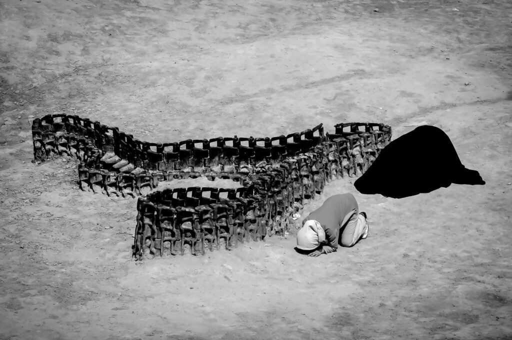 A mother and daughter kneel in mourning beside the skeletal remains of a tank, their heads bowed in prayer. The rusted carcass curves through the earth like a spine, a scar etched into the landscape by war.