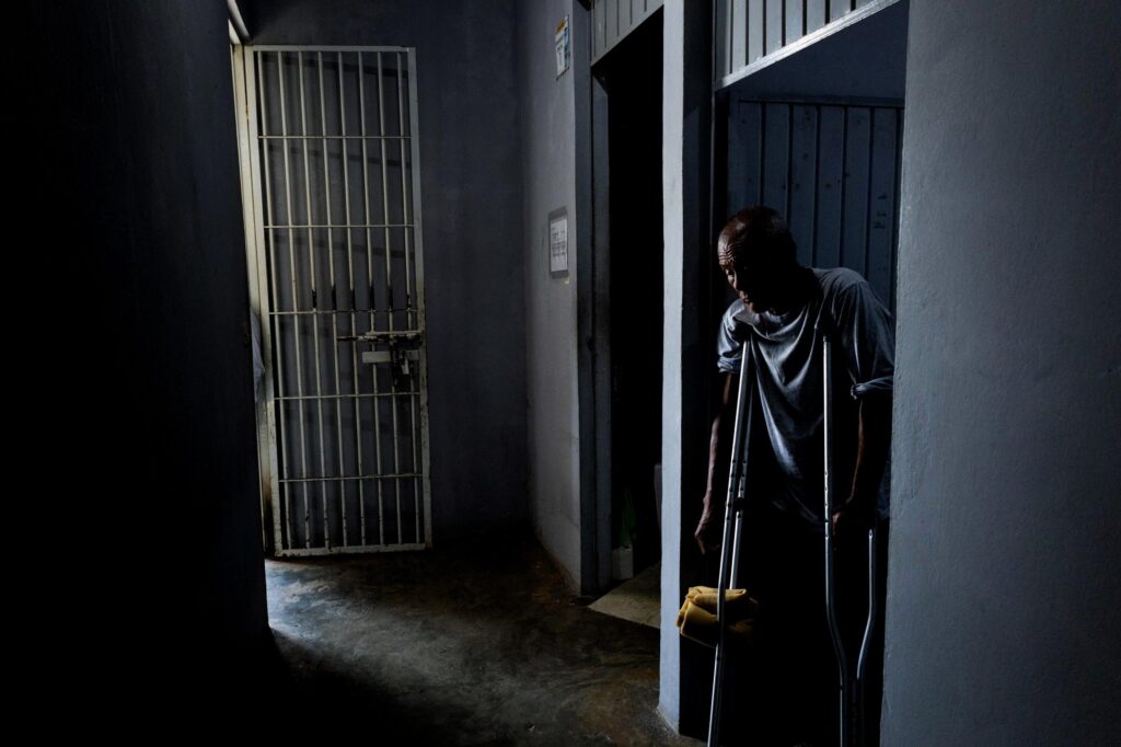 An elderly man emerges from his cell on crutches. The corridor is dim, lit only by the glare of a barred doorway, where the sleeve of a guard is just visible.