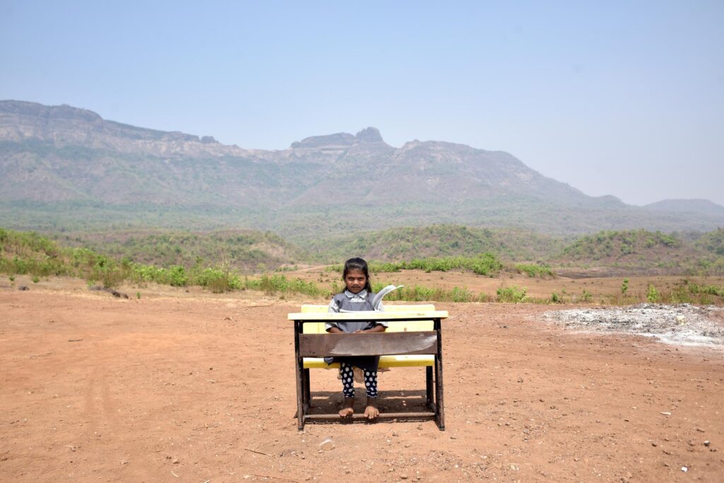 A primary school student from a tribal wadi (village) sits for a portrait outside her school, framed by the shadow of mountains and dense jungle where her community lives. Feet resting on the desk’s lower bar, hands gently clasped, she sits upright, squinting against the sun as she returns our gaze.