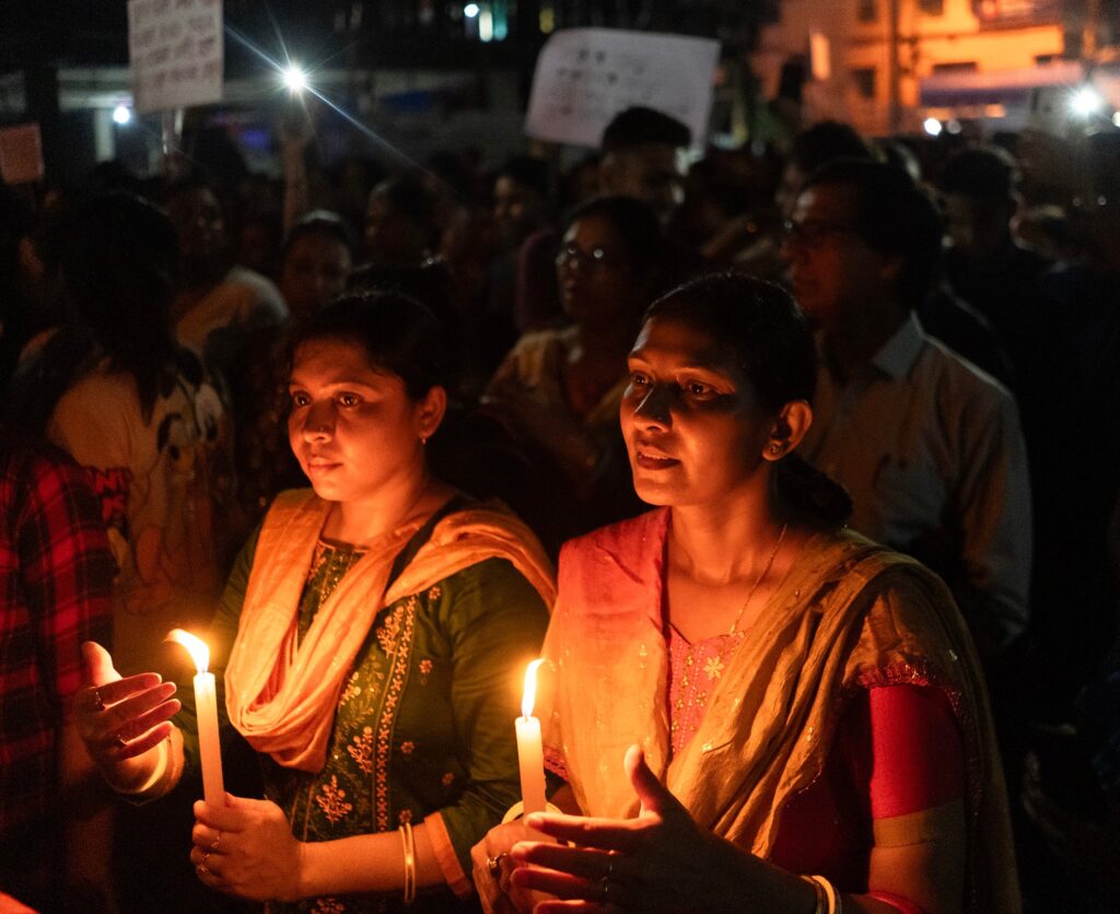 Two women stand illuminated by the candles they hold. Their faces are soft with mourning yet lit by resolve, hands curved protectively around the fragile flames. Behind them, the shadows swell with others—women gathered in protest.