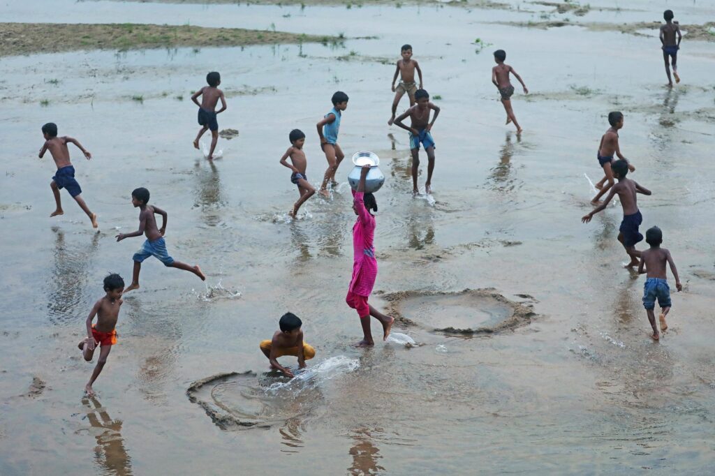 At dusk, a young girl walks home carrying water from the river as boys from her village play freely in the shallows. The scene is full of motion, the boys’ game, the shimmer of the current, the fading light, yet she moves in another rhythm, bound to the daily work that defines her place within the landscape.