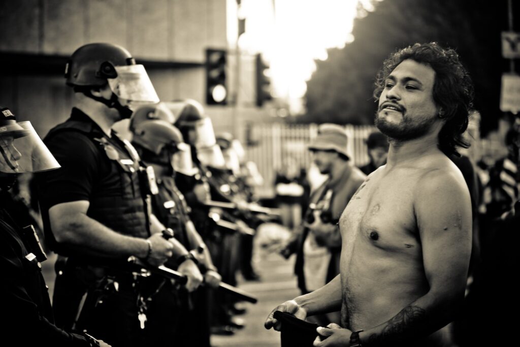 A protester stands before police during an anti-ICE demonstration in Los Angeles, baring his chest to reveal the scar left by law enforcement. He lifts his chin high in defiance, facing the armoured line without fear. His body becomes both evidence and resistance, a counterpoint to their riot shields.