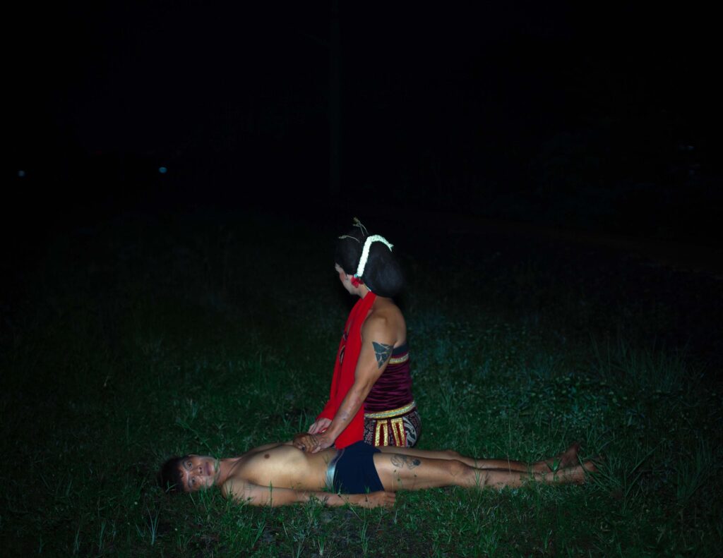 One body rests in the grass, facing the camera, while another kneels above in ceremonial attire inspired by Lengger, a Javanese cross-dressing dance. Set against the darkness of night, the scene evokes a quiet ritual of tenderness and healing.