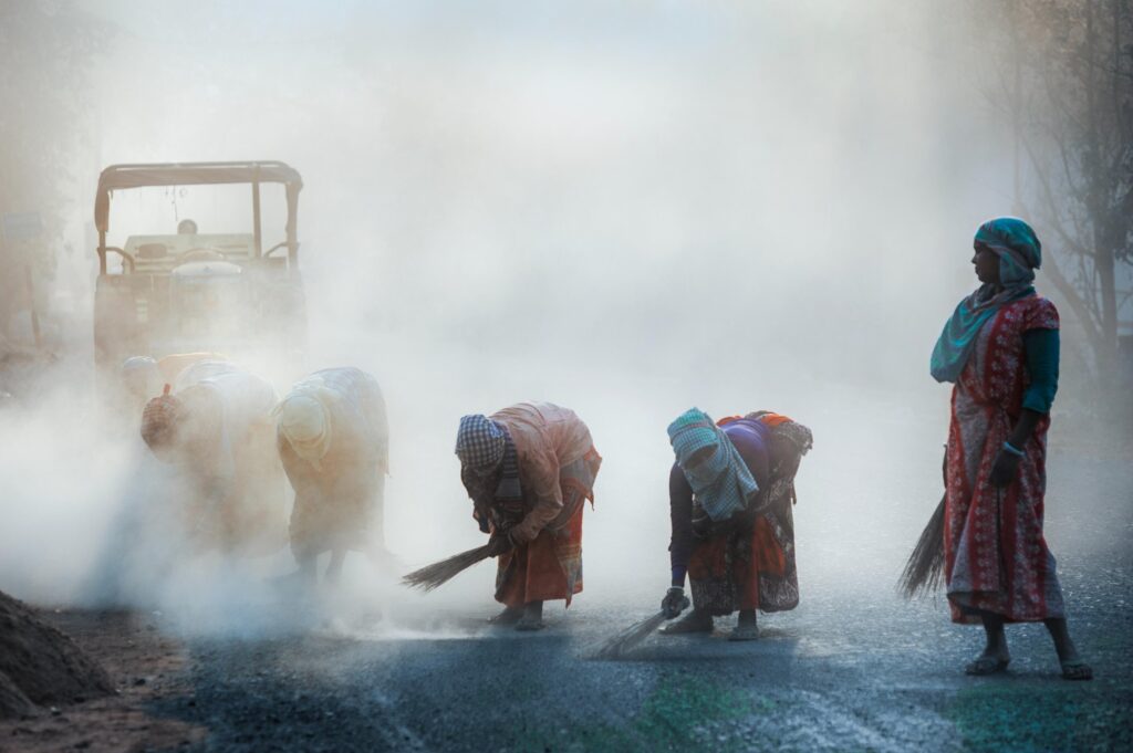 Dust rises in the air as a group of women repair a road in Bangladesh. Bent over their brooms, they work through the haze; one woman pauses, broom in hand, watching over the others.
